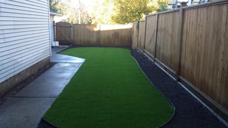 Installed artificial turf lawn in a narrow backyard space next to a concrete path and wood fence.